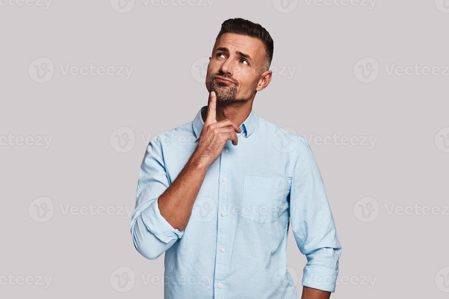 Looking for fresh ideas. Thoughtful young man keeping hand on chin and looking away while standing against grey background photo