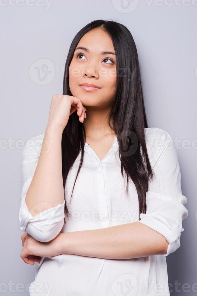 Lost in thoughts. Thoughtful young Asian woman holding hand on chin and looking away while standing grey background photo
