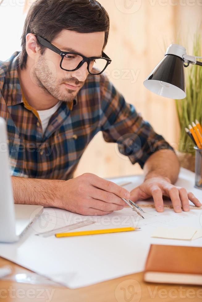 Turning his ideas to reality. Concentrated young man using compass for drawing while sitting at his workplace photo