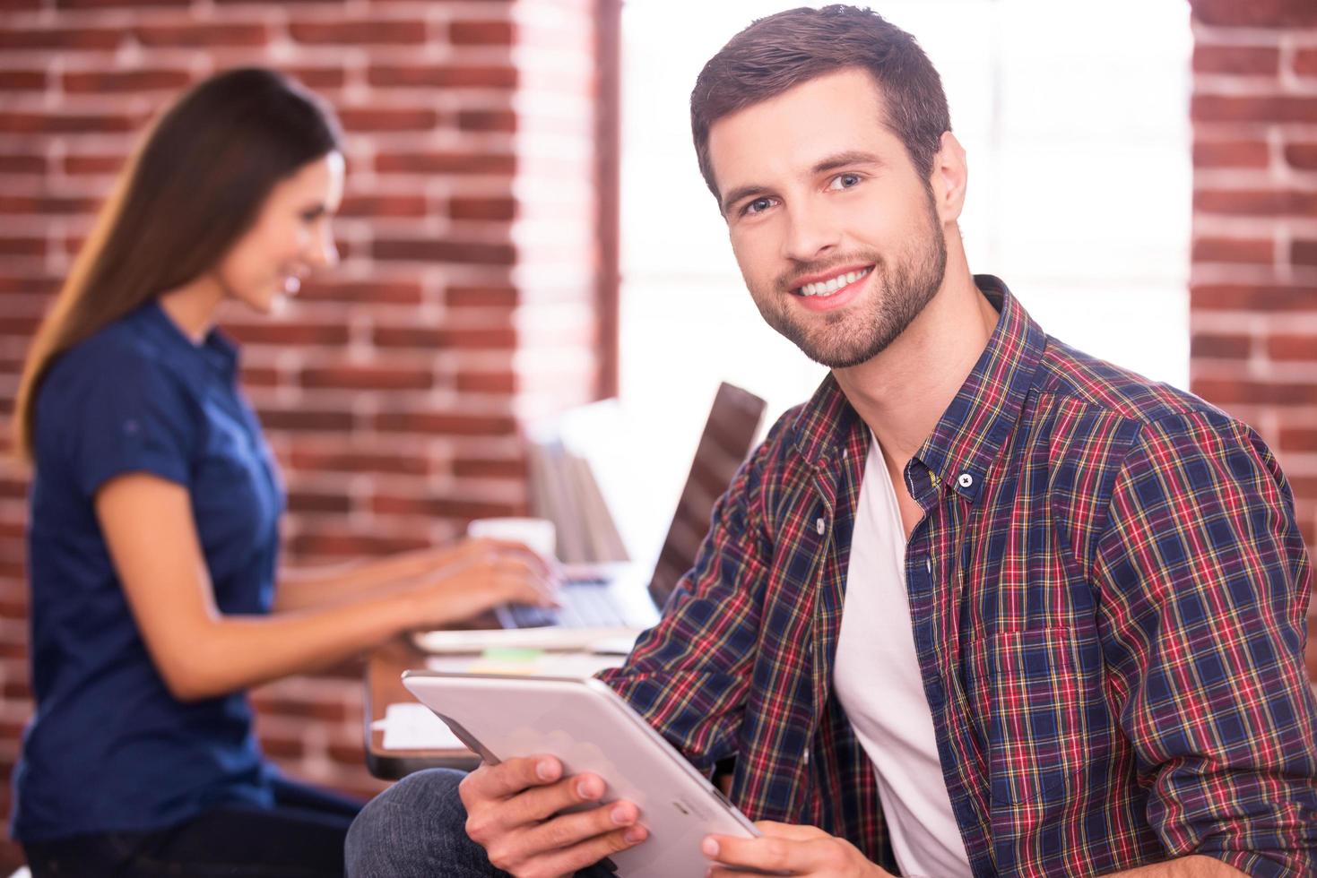Confident and creative. Handsome young man holding digital tablet and smiling while woman sitting in the background and working on laptop photo