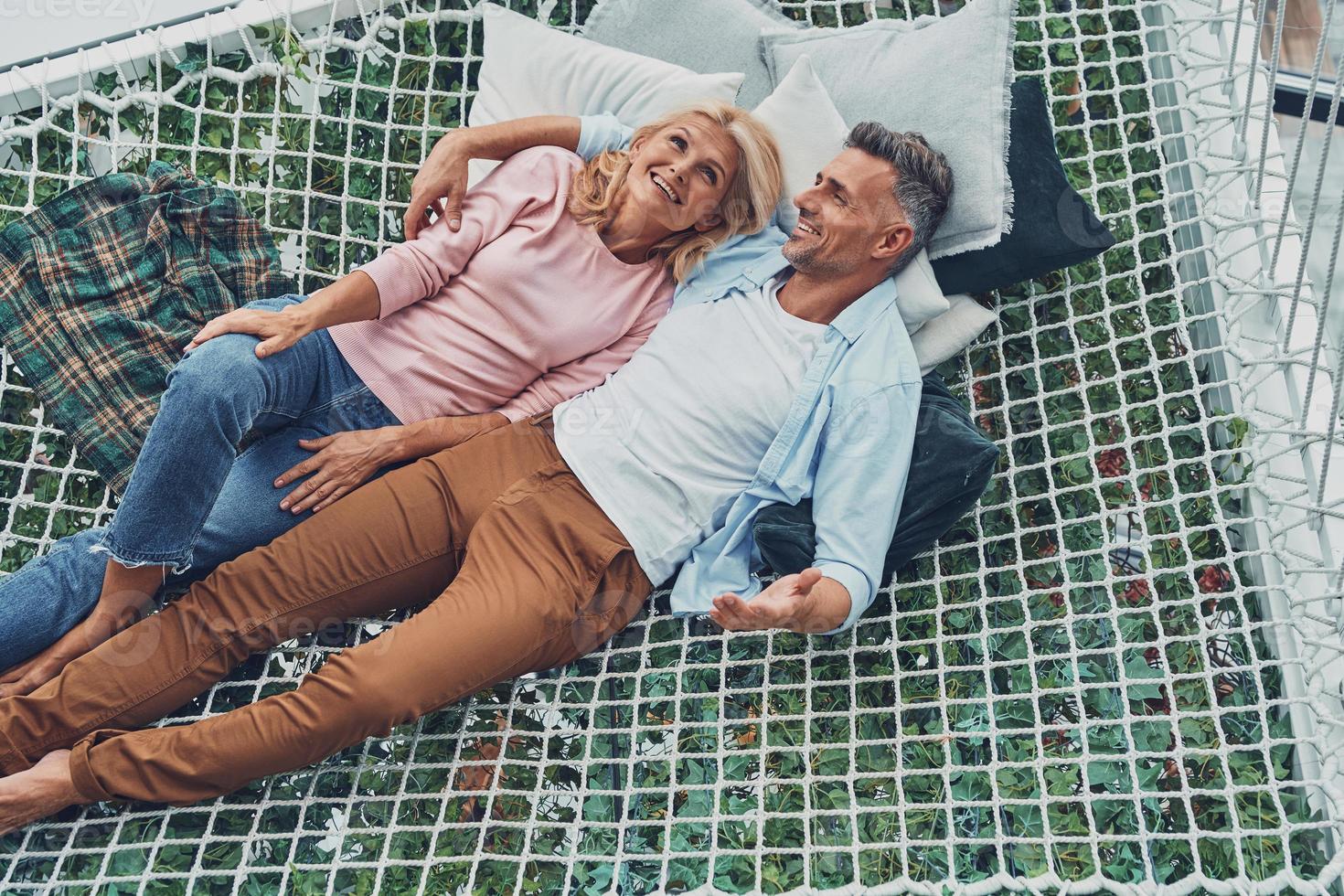 Top view of beautiful mature couple smiling and communicating while relaxing in big hammock at home together photo