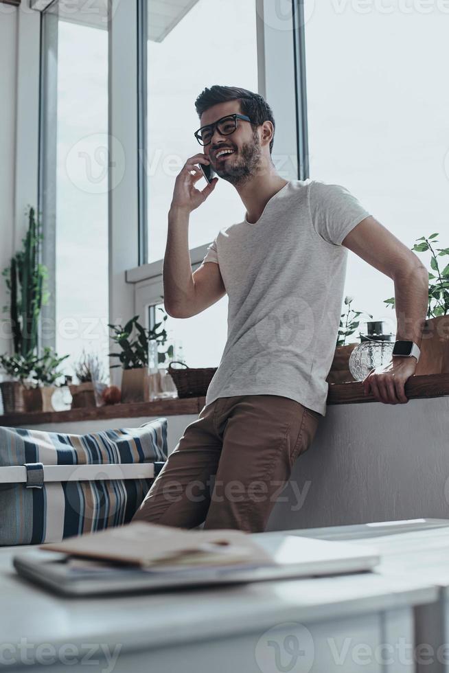Always in touch with friends. Handsome young man talking on his smart phone while leaning on the window sill photo