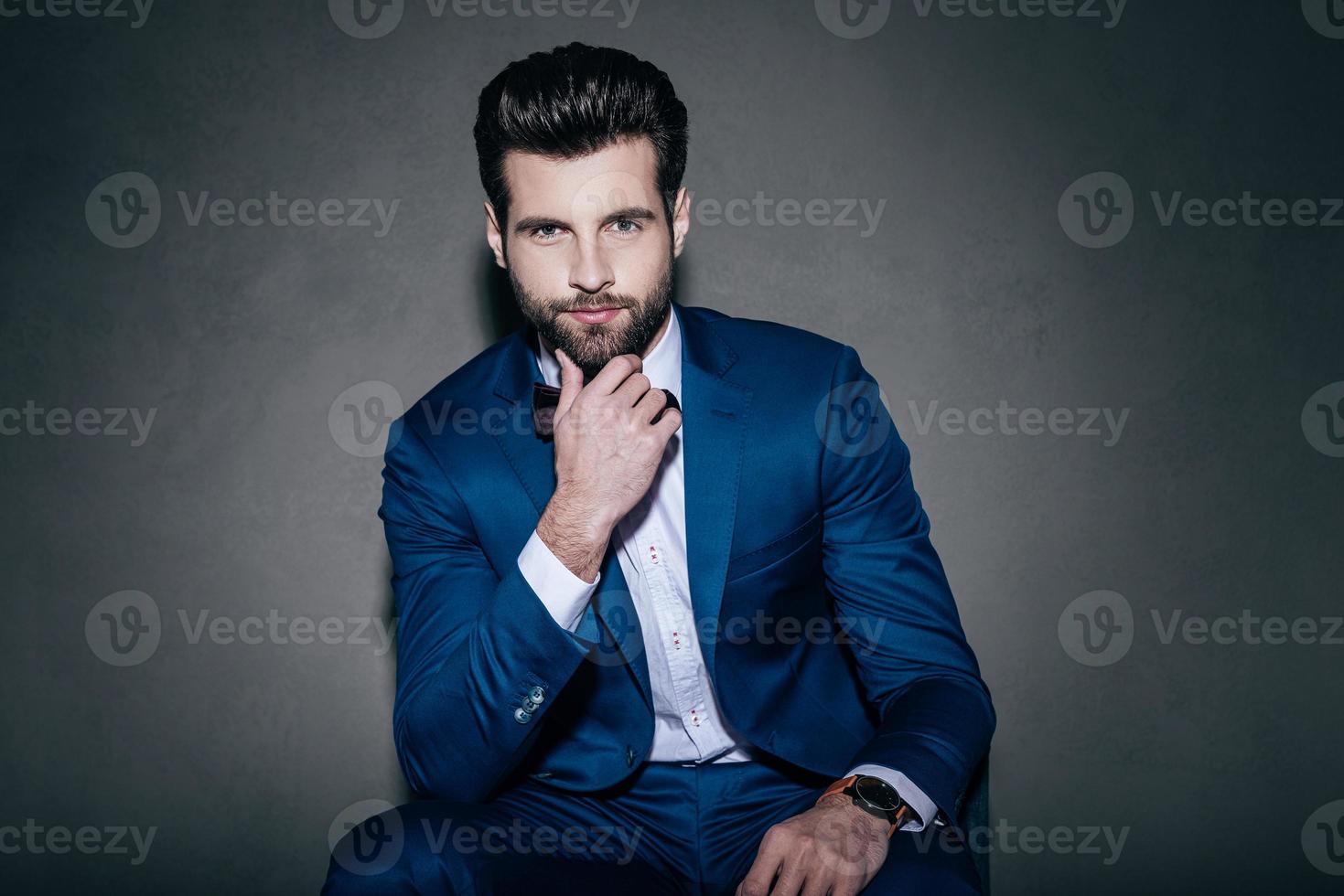 perfecto guapo. joven apuesto con traje y corbata de moño mirando a la cámara y sosteniendo la mano en la barbilla mientras se sienta en un fondo gris foto