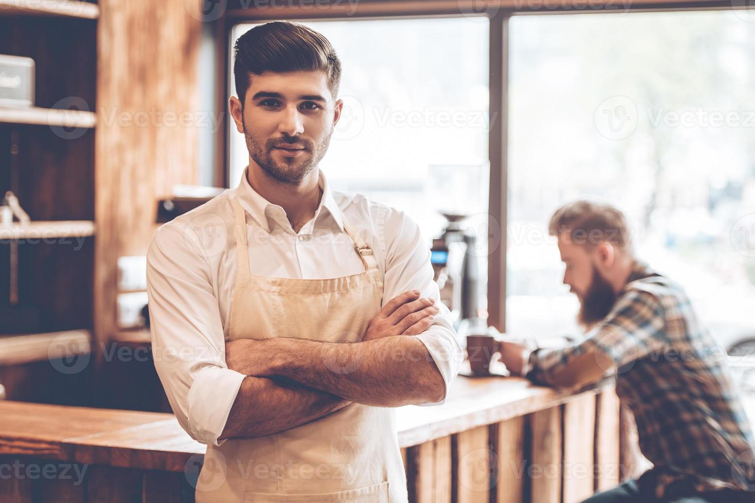 Loving his work. Young handsome man in apron looking at camera and keeping arms crossed while standing at cafe with customer at background photo