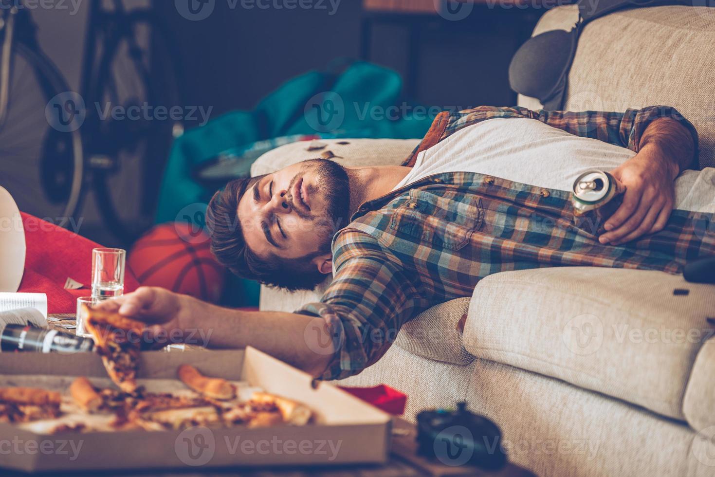 It has been a long night. Young handsome man passed out on sofa with pizza slice and beer can in his hand in messy room after party photo