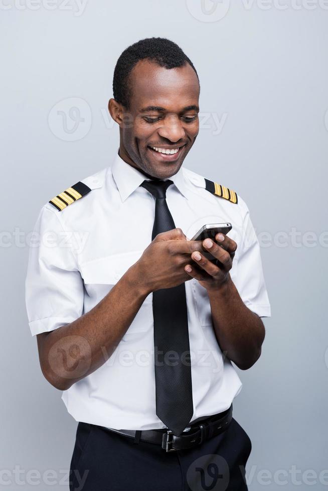 Messages after landing. Side view of happy African pilot in uniform holding mobile phone while standing against grey background photo