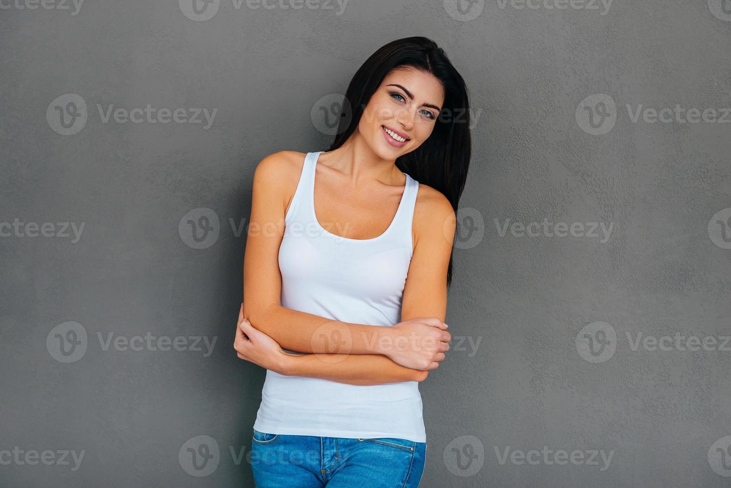 Elegance and beauty. Beautiful young woman in white tank top keeping arms crossed and smiling while standing against grey background photo