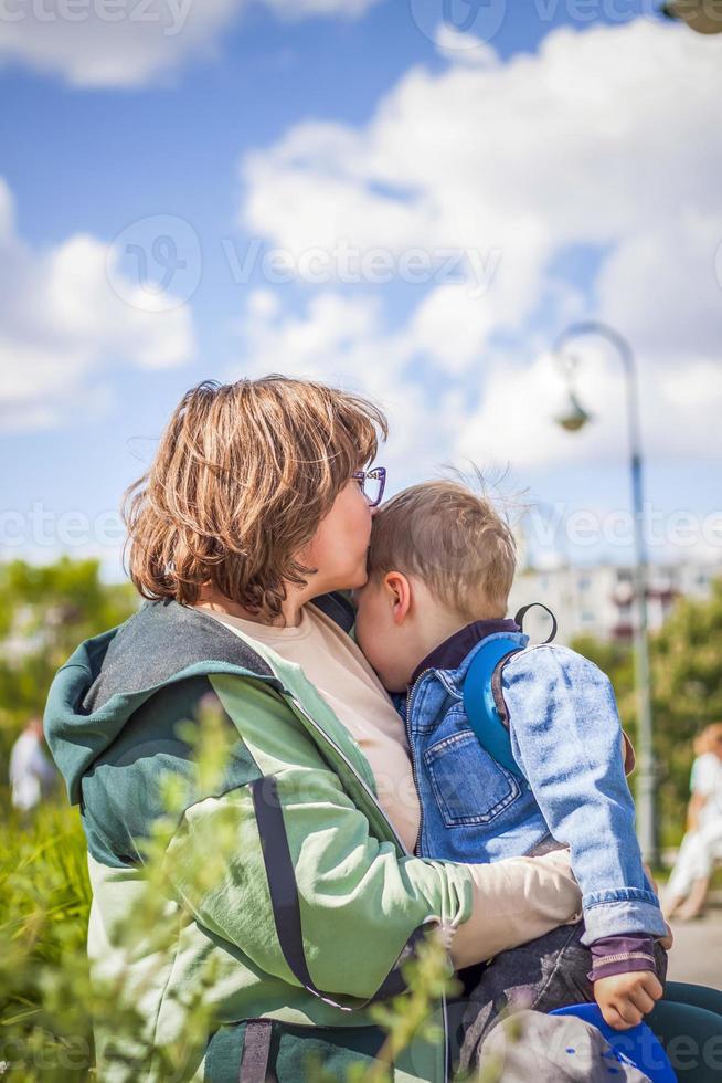 A moody child cuddles up to his young grandmother in the park. The woman feels sorry for the boy in her arms. Interactions. Selective focus. photo