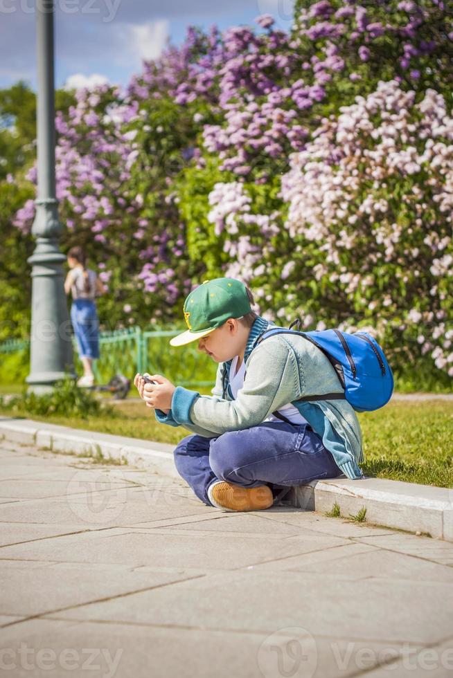 un joven está sentado al lado de un callejón lila y mira su teléfono. contra el fondo de arbustos de lilas. interacciones. enfoque selectivo. foto