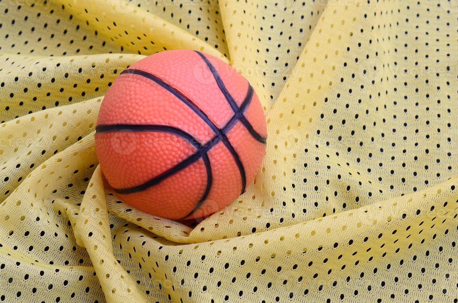 Small orange rubber basketball lies on a yellow sport jersey clothing fabric texture and background with many folds photo