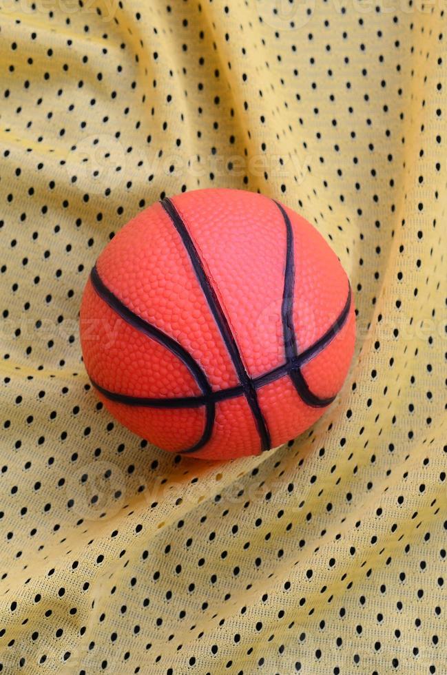 Small orange rubber basketball lies on a yellow sport jersey clothing fabric texture and background with many folds photo