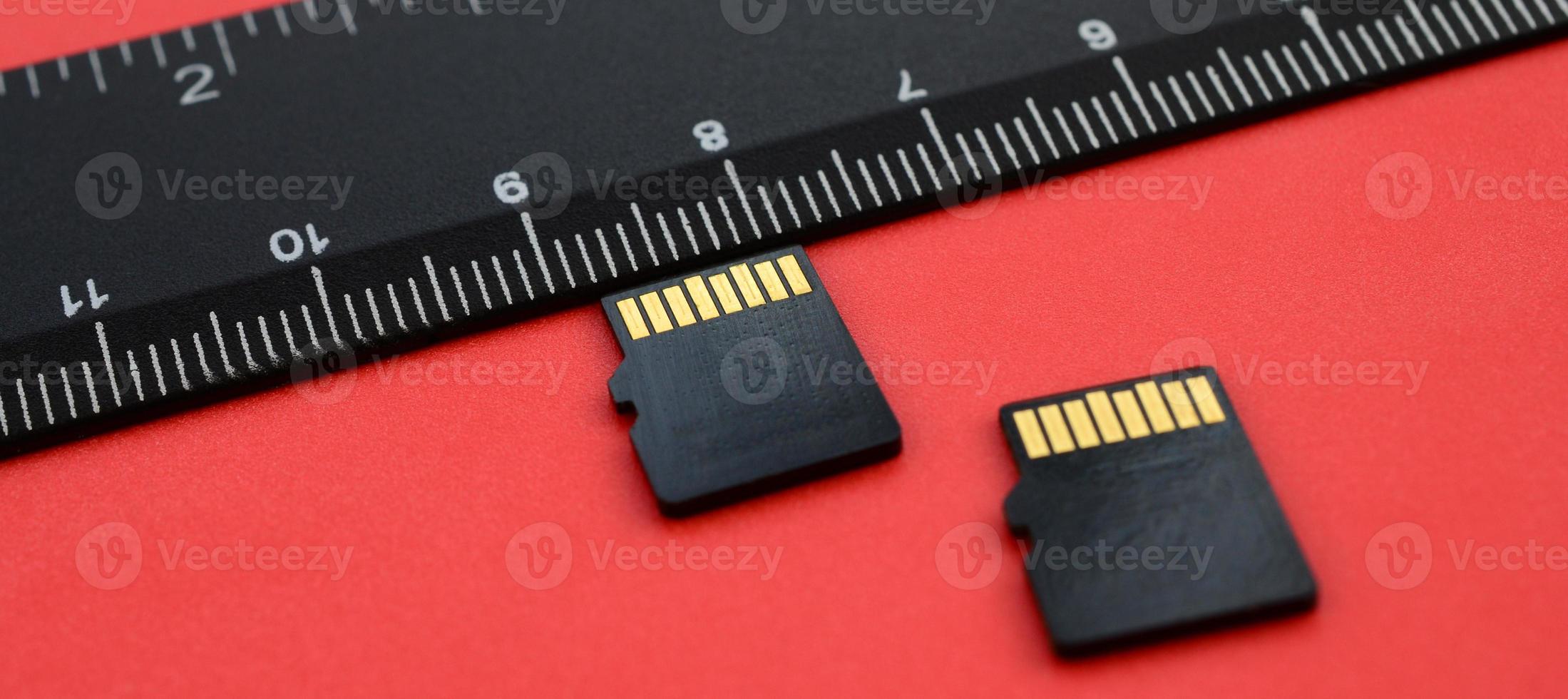 Two small micro SD memory cards lie on a red background next to a black ruler. A small and compact data and information store photo