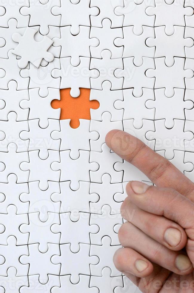 The texture of a white jigsaw puzzle in the assembled state with one missing element, forming an orange space, pointed to by the finger of the male hand photo