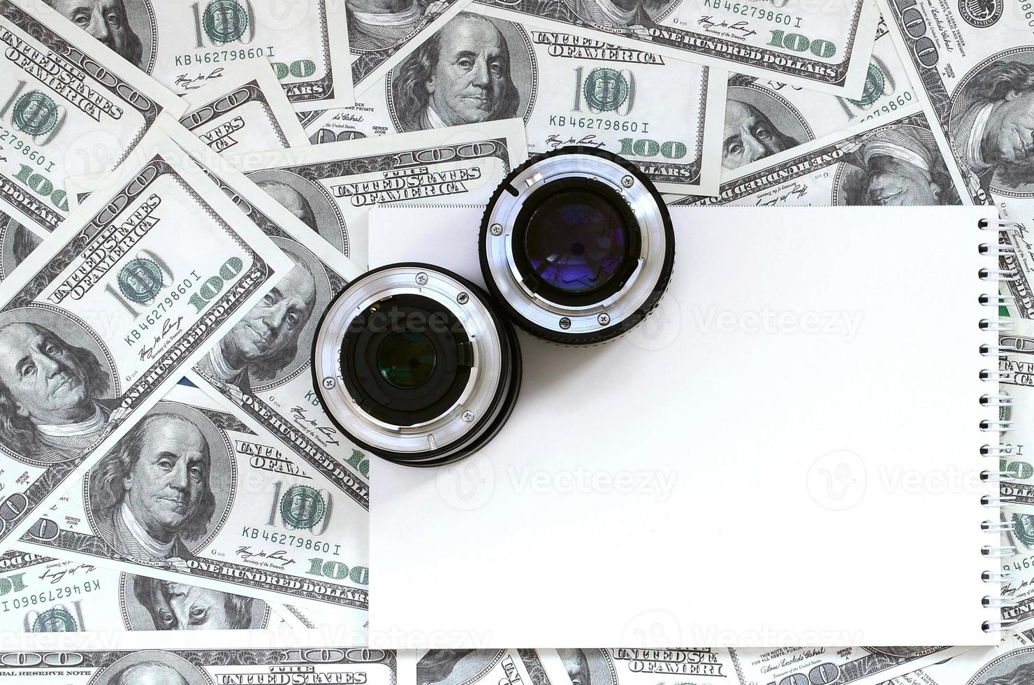 Two photographic lenses and white notebook lie on the background of a lot of dollar bills. Space for text photo