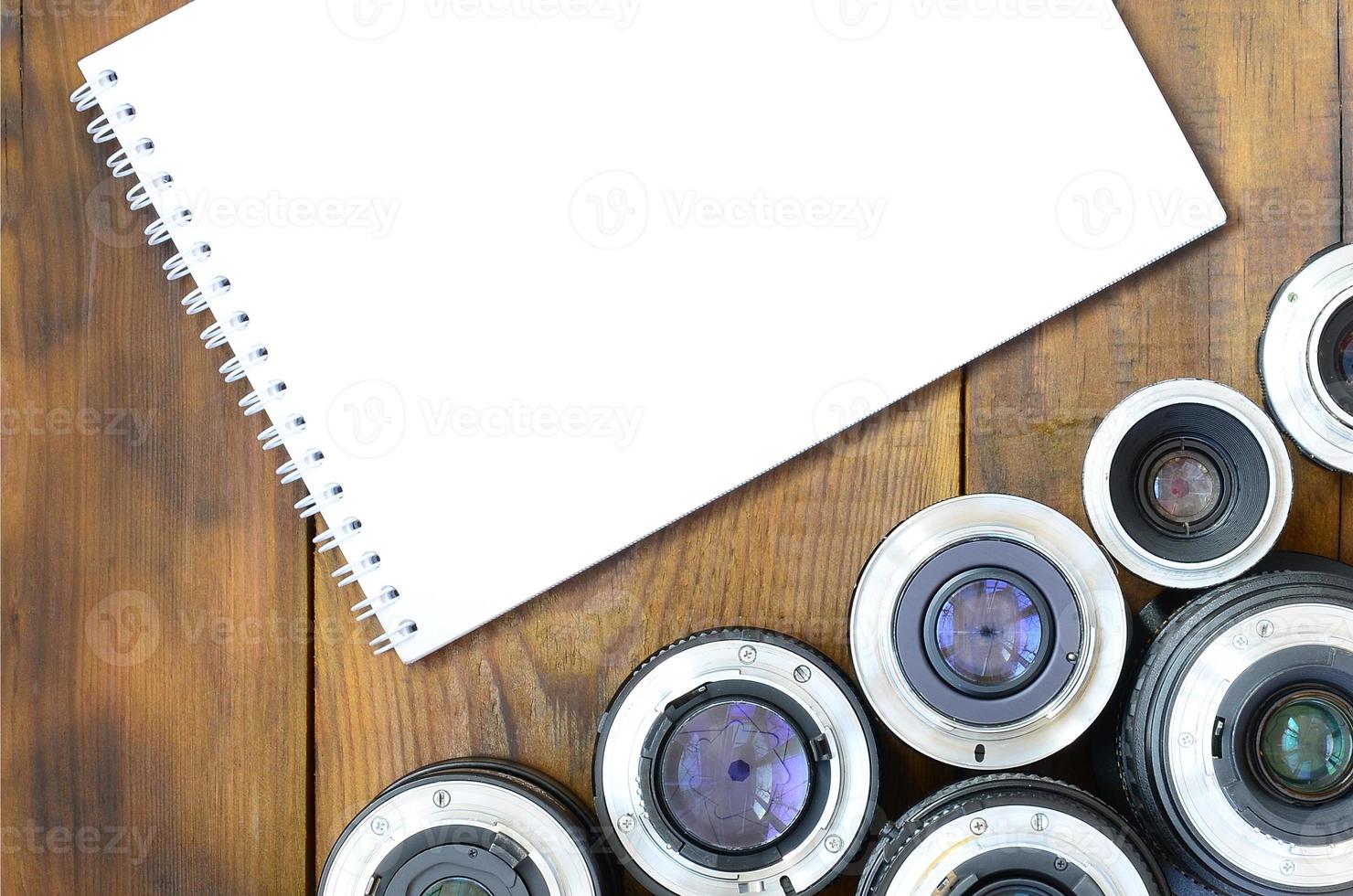 Several photographic lenses and white notebook lie on a brown wooden background. Space for text photo