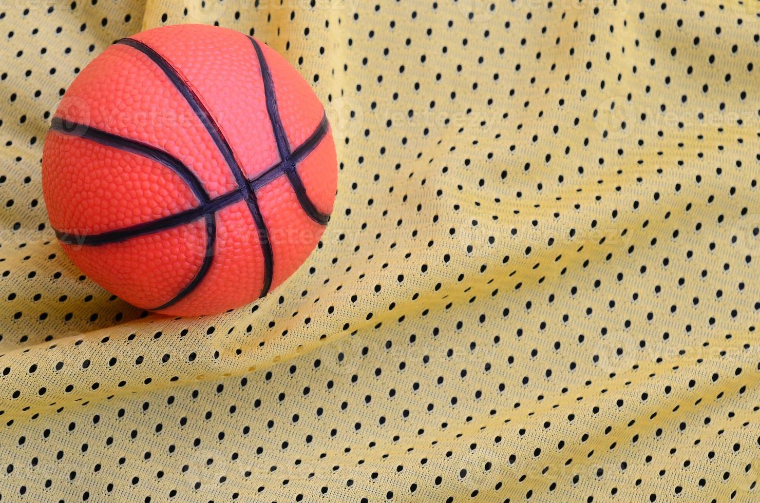 Small orange rubber basketball lies on a yellow sport jersey clothing fabric texture and background with many folds photo