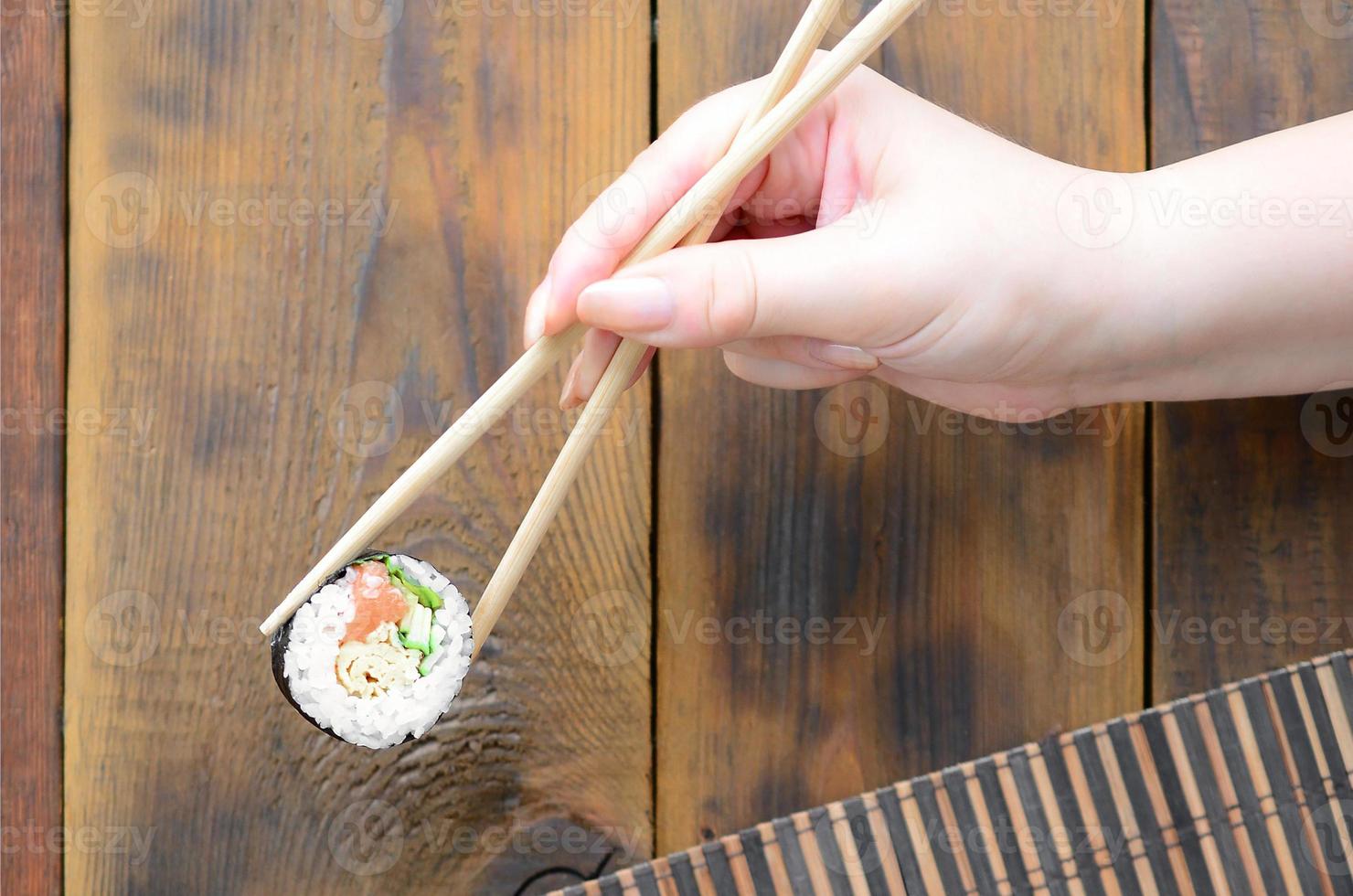 A hand with chopsticks holds a sushi roll on a bamboo straw serwing mat background. Traditional Asian food photo