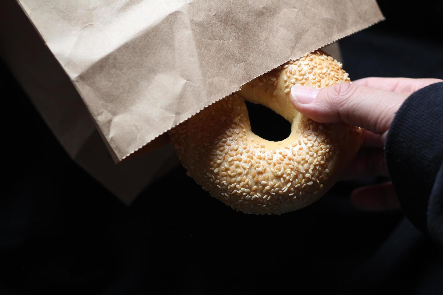 A man's hand is holding a bagel out of a brown paper bag. photo