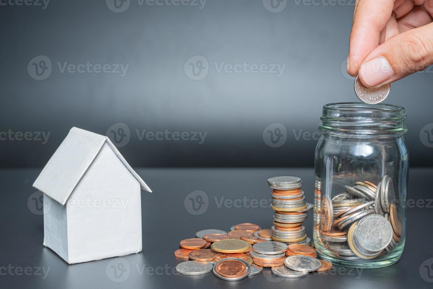 Hand putting Thailand coins in jar with heap coins on black background with house model photo