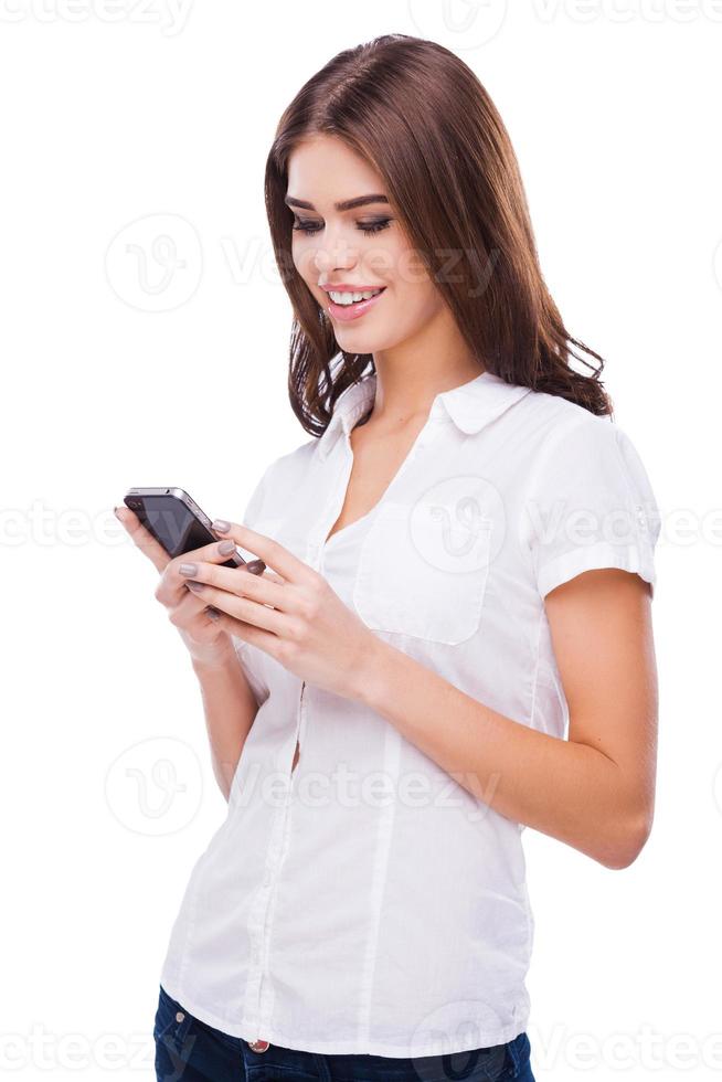 Sending message to him. Beautiful young women holding mobile phone and smiling while standing against white background photo