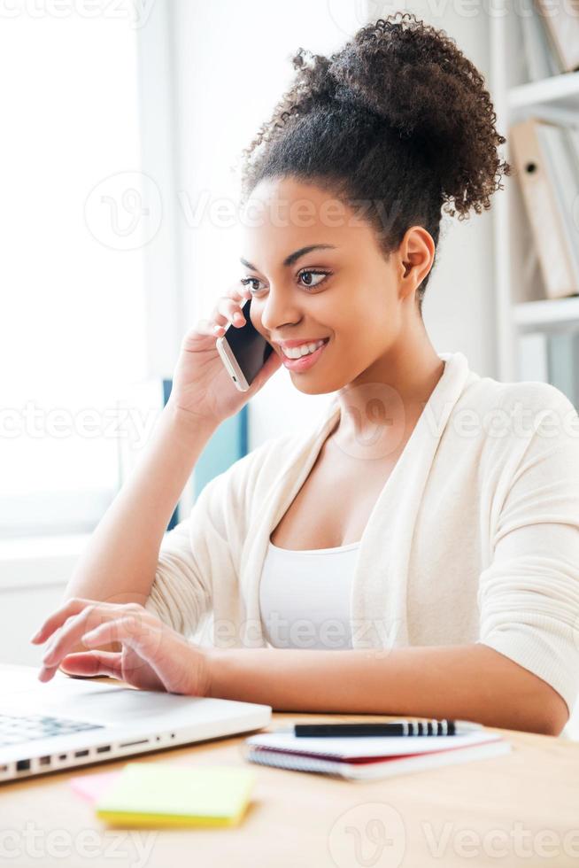 trabajando con placer. bella joven africana hablando por teléfono móvil y trabajando en una laptop mientras se sienta en su lugar de trabajo en la oficina foto