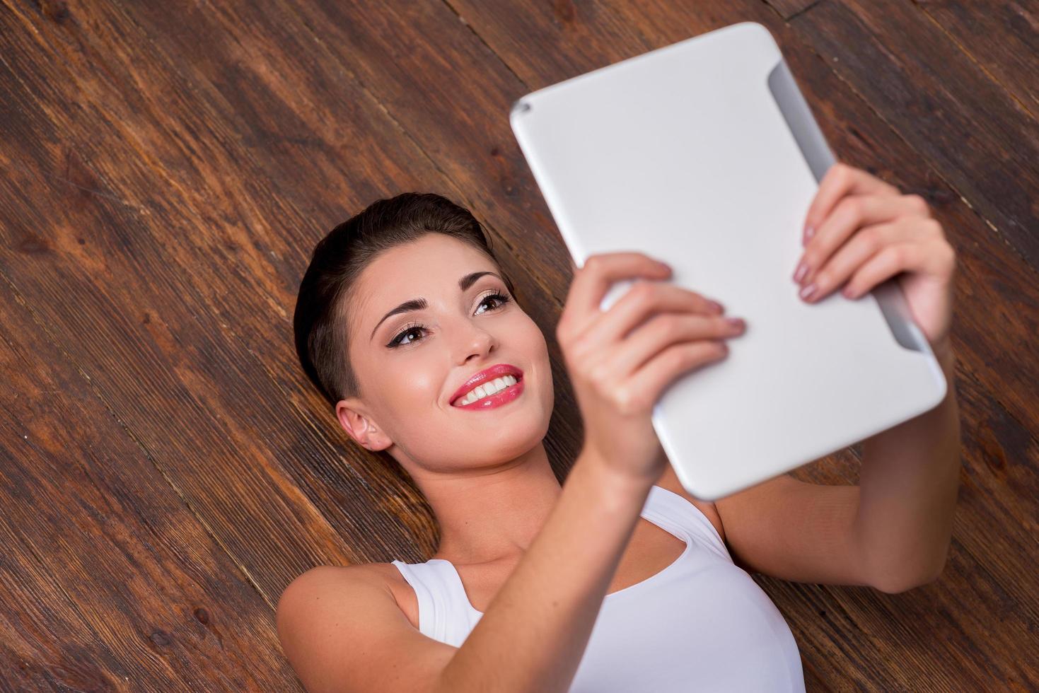 Relaxing with her brand new tablet. Top view of beautiful young short hair woman holding digital tablet and smiling while lying on the hardwood floor photo