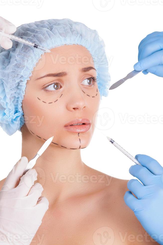Handmade beauty. Scared young woman keeping looking away while four hands in medical gloves holding syringes and knifes close to her face photo