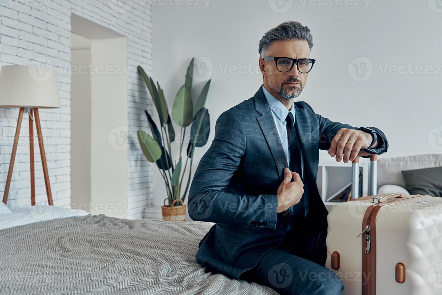 Handsome man in formalwear leaning on his luggage and looking at camera while sitting on bed photo