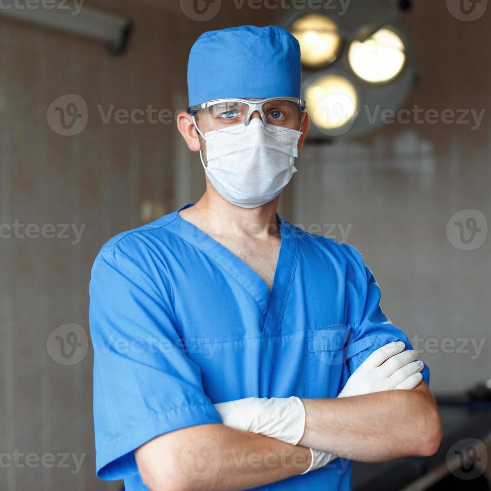cirujano con uniforme azul, gafas y sombreros en el quirófano al fondo de luces brillantes foto