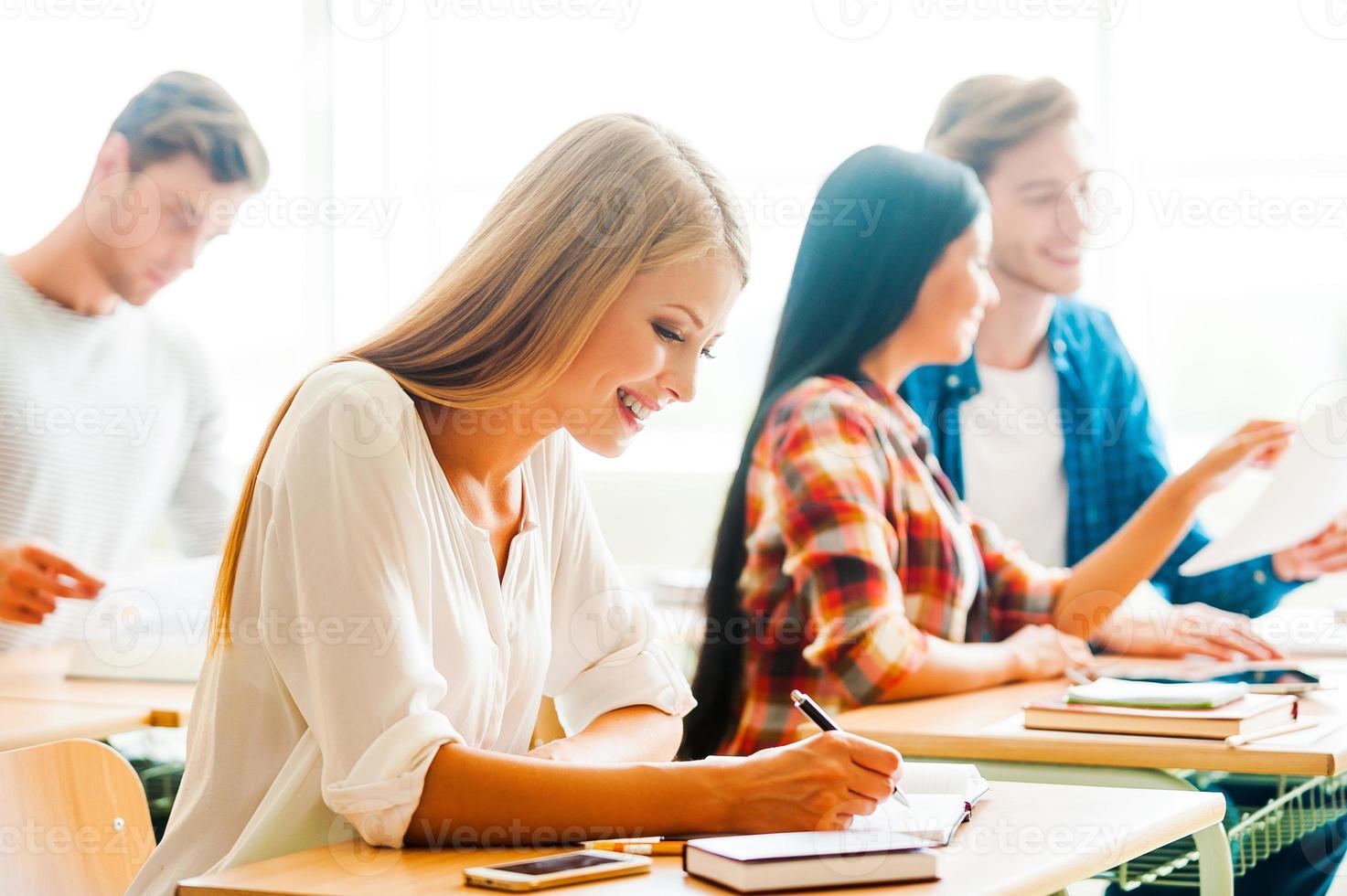 Studying with pleasure. Happy young woman writing in note pad and smiling while sitting at her desk in the classroom photo