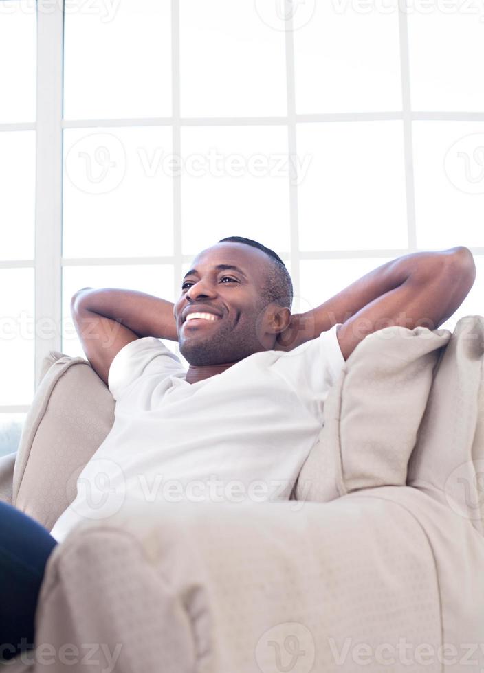 Relaxing at home. Cheerful African man holding head in hands and smiling while sitting on the chair photo