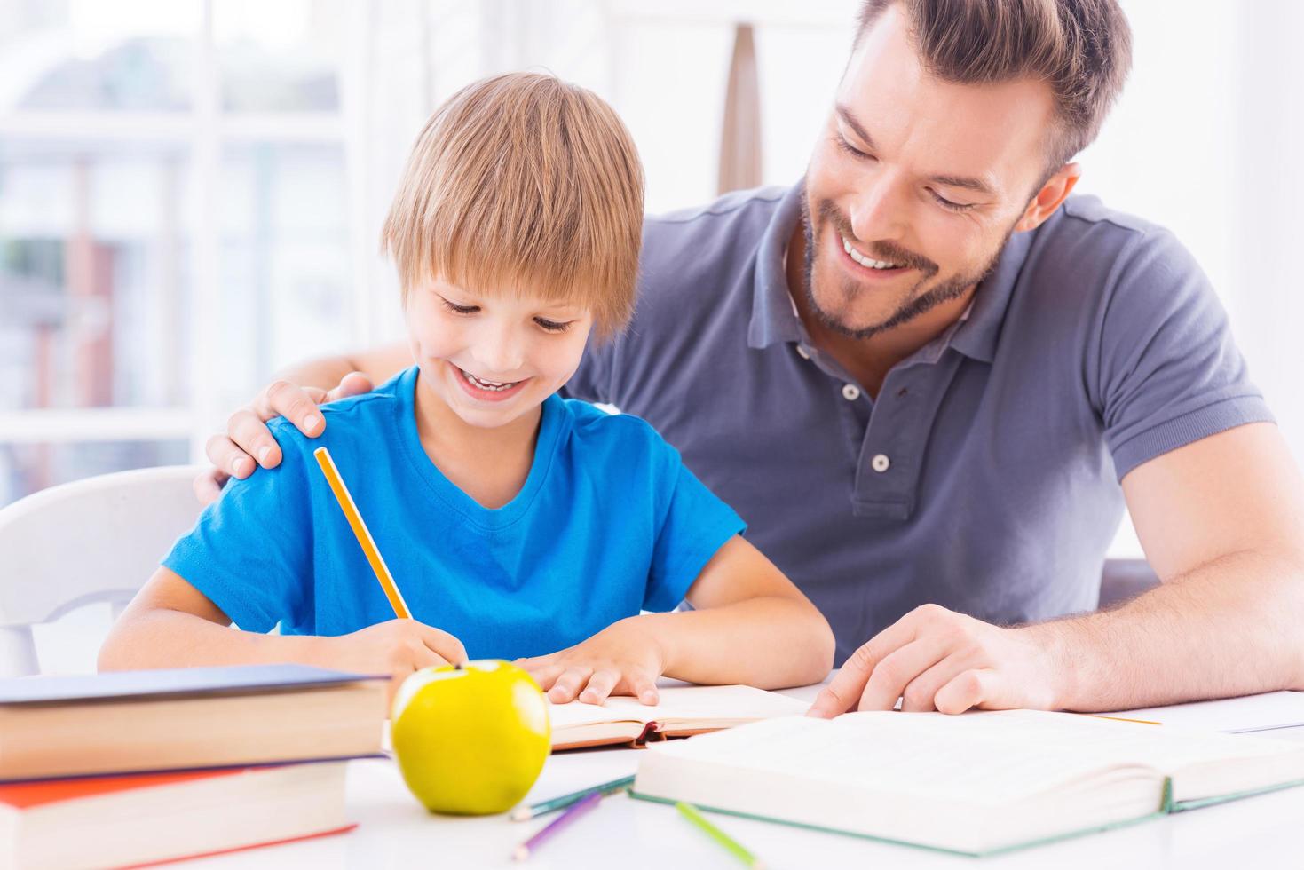 ayudar a hijo con el trabajo escolar. alegre padre joven ayudando a su hijo con la tarea y sonriendo mientras se sientan juntos en la mesa foto