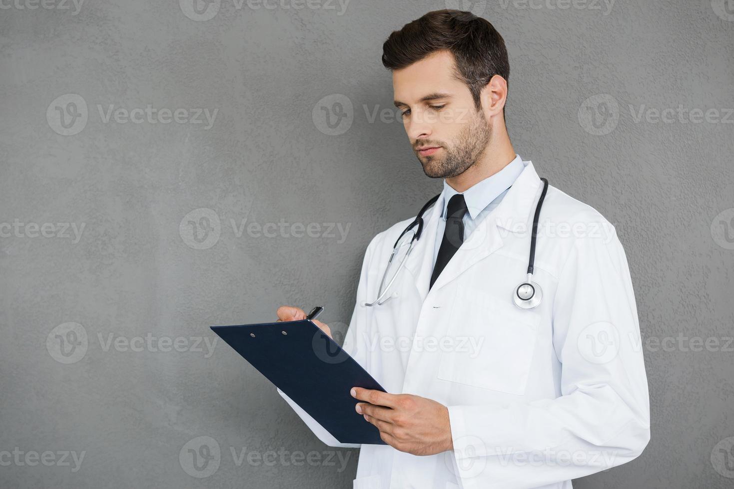 Writing the prescription. Confident young doctor in white uniform writing in clipboard while standing against grey background photo