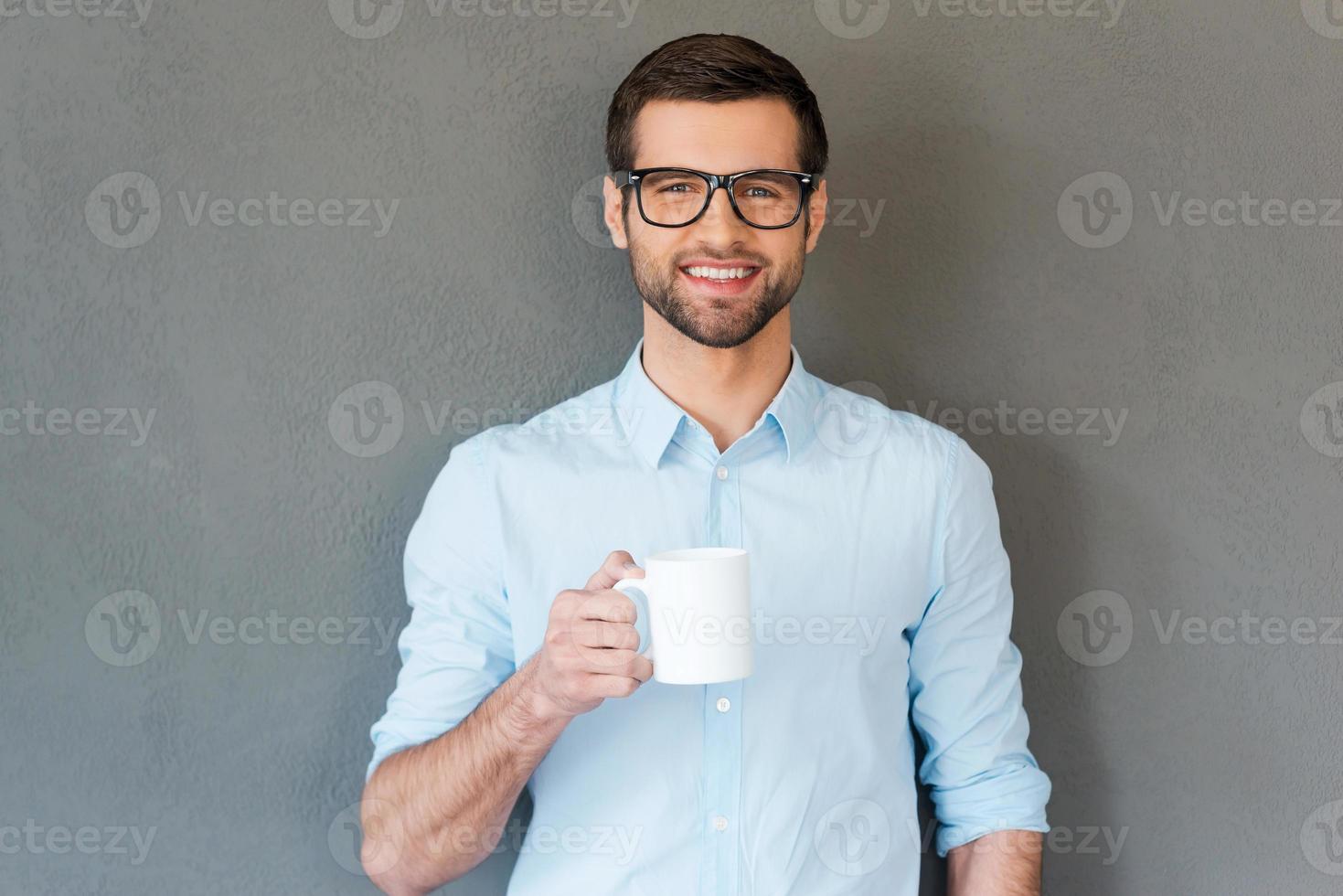 Good coffee for starting your day. Handsome young man in shirt in eyewear holding cup of coffee and smiling at camera while standing against grey background photo