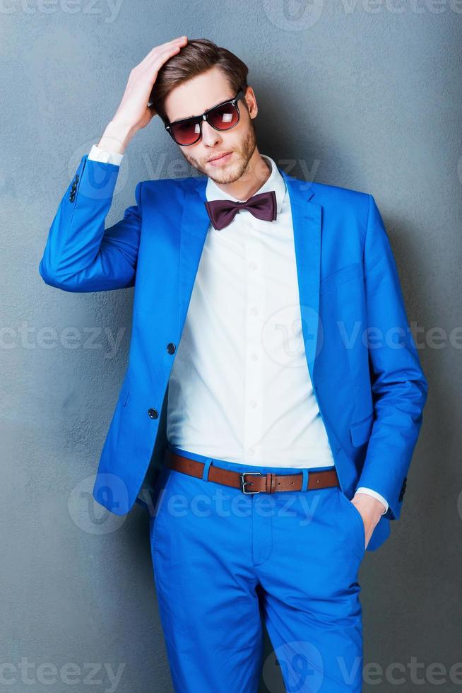He knows how to be stylish. Serious young man in eyewear holding hand in hair and looking at camera while standing against grey background photo