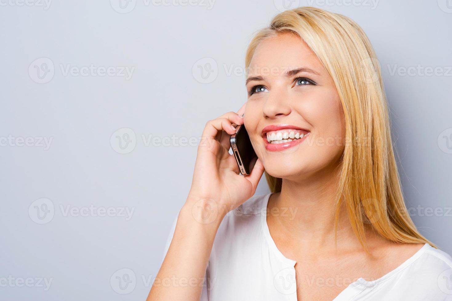 Phone emotions. Beautiful young women talking on the mobile phone and smiling while standing against grey background photo