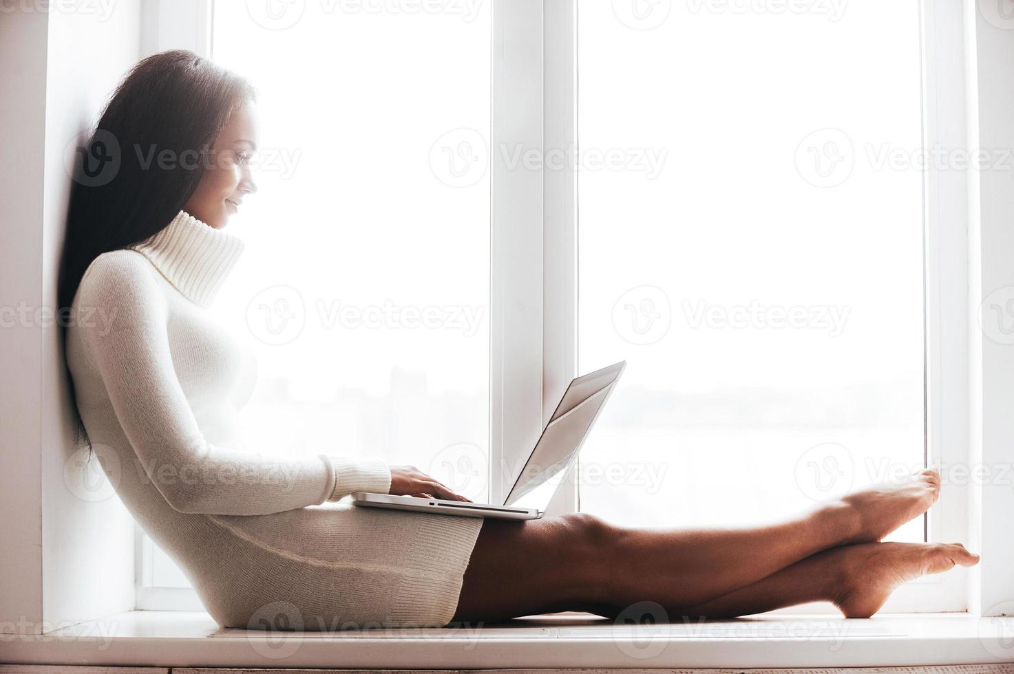 Beauty with laptop. Attractive young African woman in warm sweater working on laptop while sitting on the window sill photo