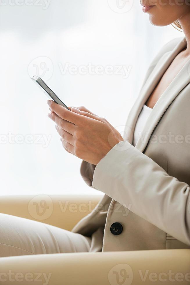Typing business message. Side view of businesswoman in suit typing something on mobile phone while sitting at the chair photo