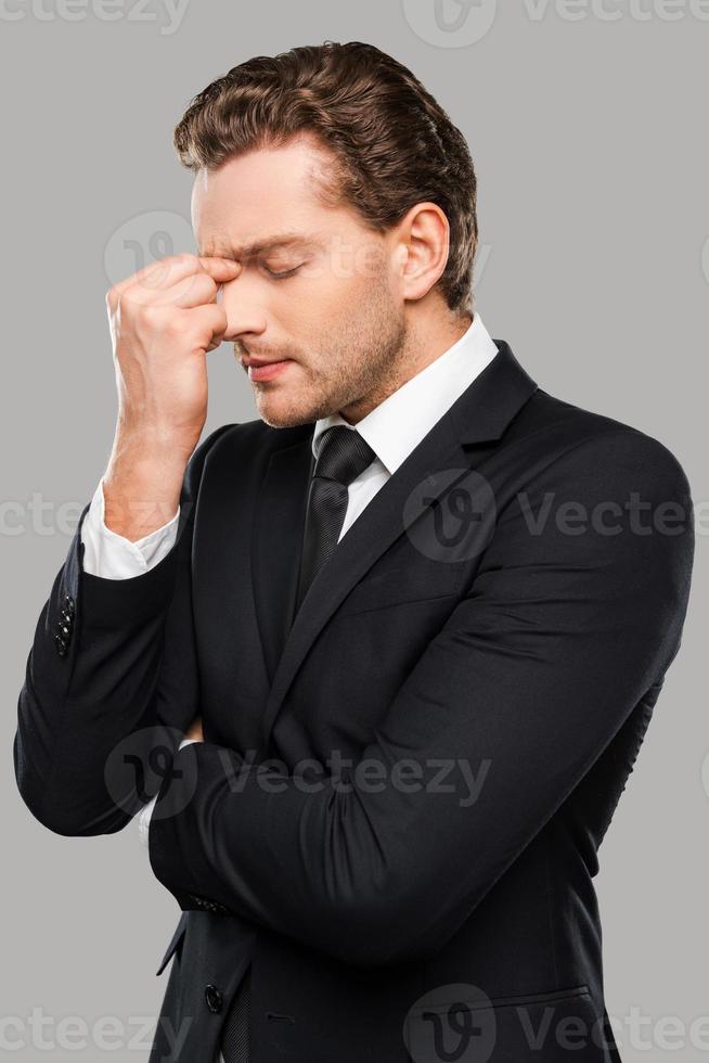 Too stressful day. Frustrated young man in formalwear touching head with fingers and keeping eyes closed while standing against grey background photo