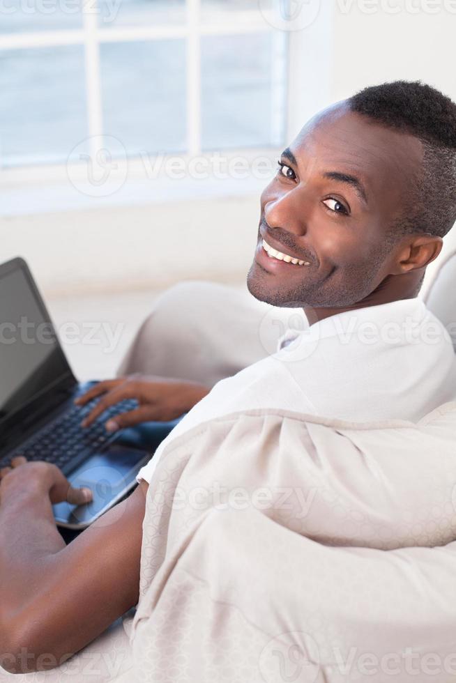 Working on laptop. Top view of  cheerful African man using computer and looking over shoulder while sitting on the chair photo