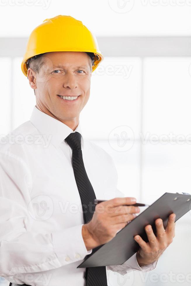 Construction foreman. Cheerful mature man in formalwear and hardhat writing something in his note pad and smiling at camera photo