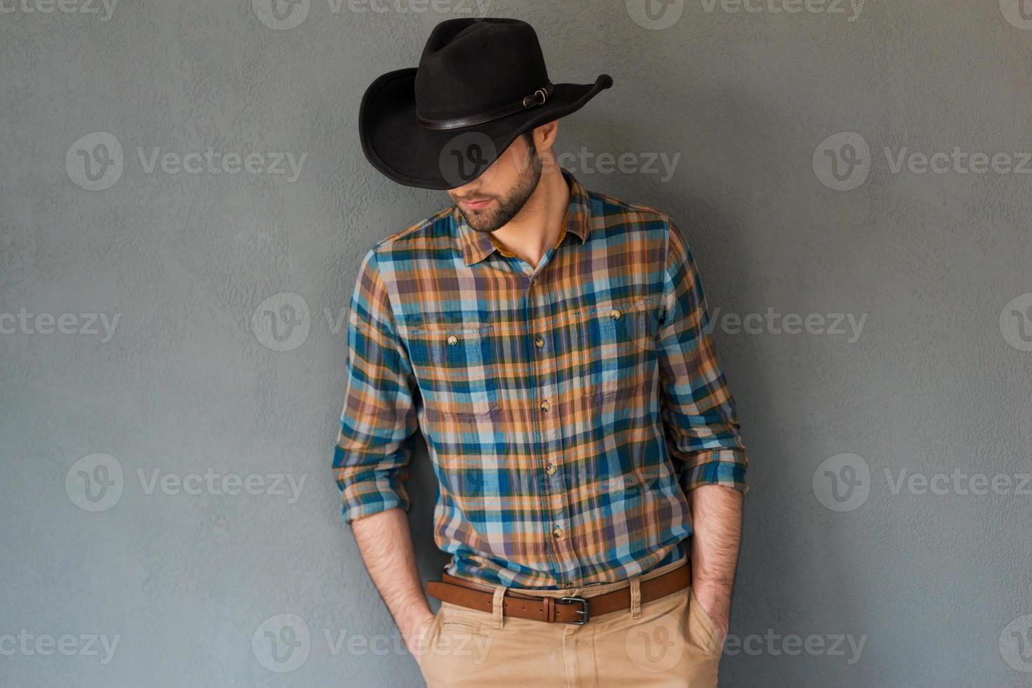 alta costura vaquera. retrato de un joven con sombrero de vaquero y mirando hacia abajo mientras se enfrenta a un fondo gris foto