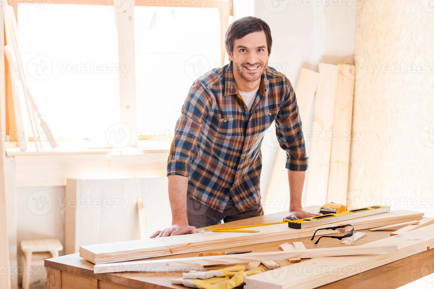 Smiling woodworker. Cheerful young male carpenter leaning at the wooden table with diverse working tools laying on it photo