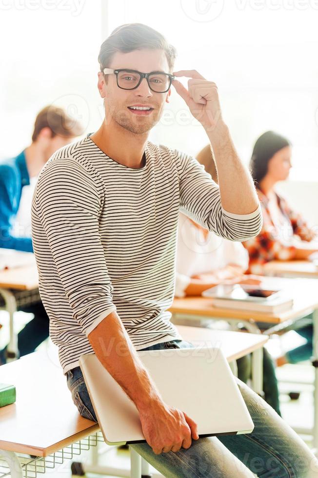 Confident student. Smiling young man holding laptop and adjusting eyewear while leaning at the desk photo