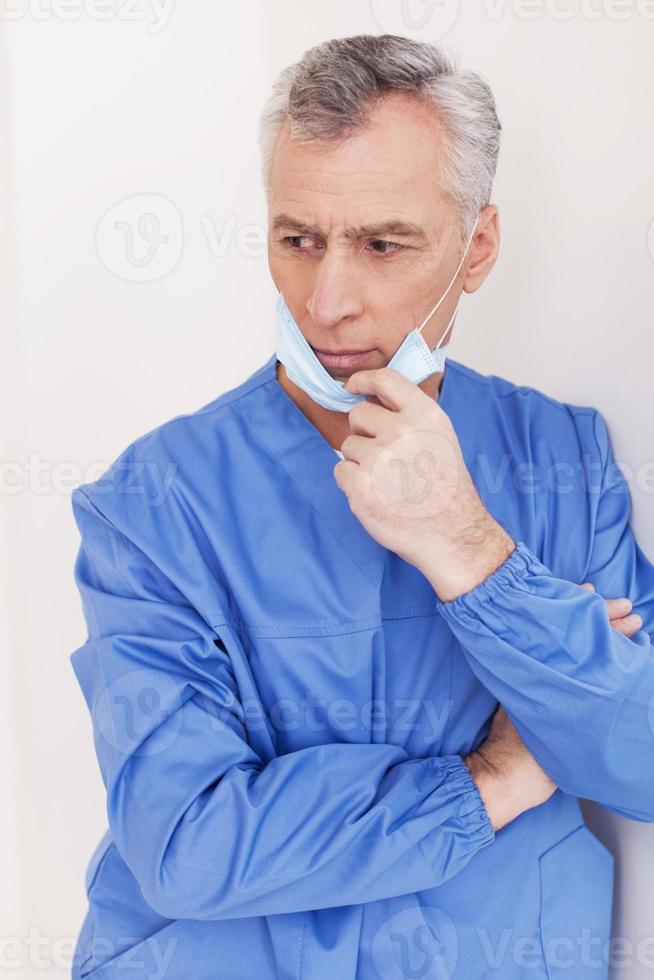 Thoughtful surgeon. Depressed senior grey hair doctor taking off his surgical mask while leaning at the wall photo