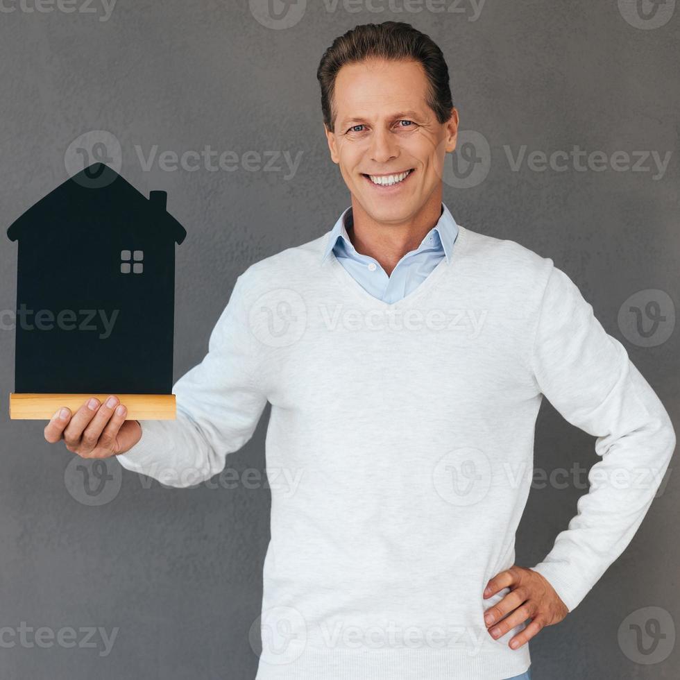 Happy house owner. Confident mature man holding house shaped object in hand and smiling while standing against grey background photo