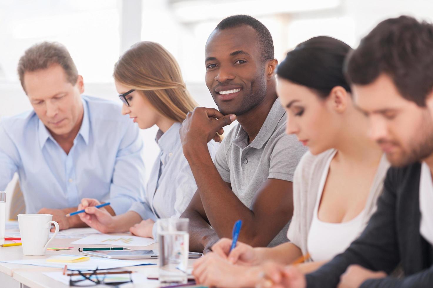 I am a part of strong and creative team. Group of business people sitting in a row at the table while handsome African man looking at camera and smiling photo