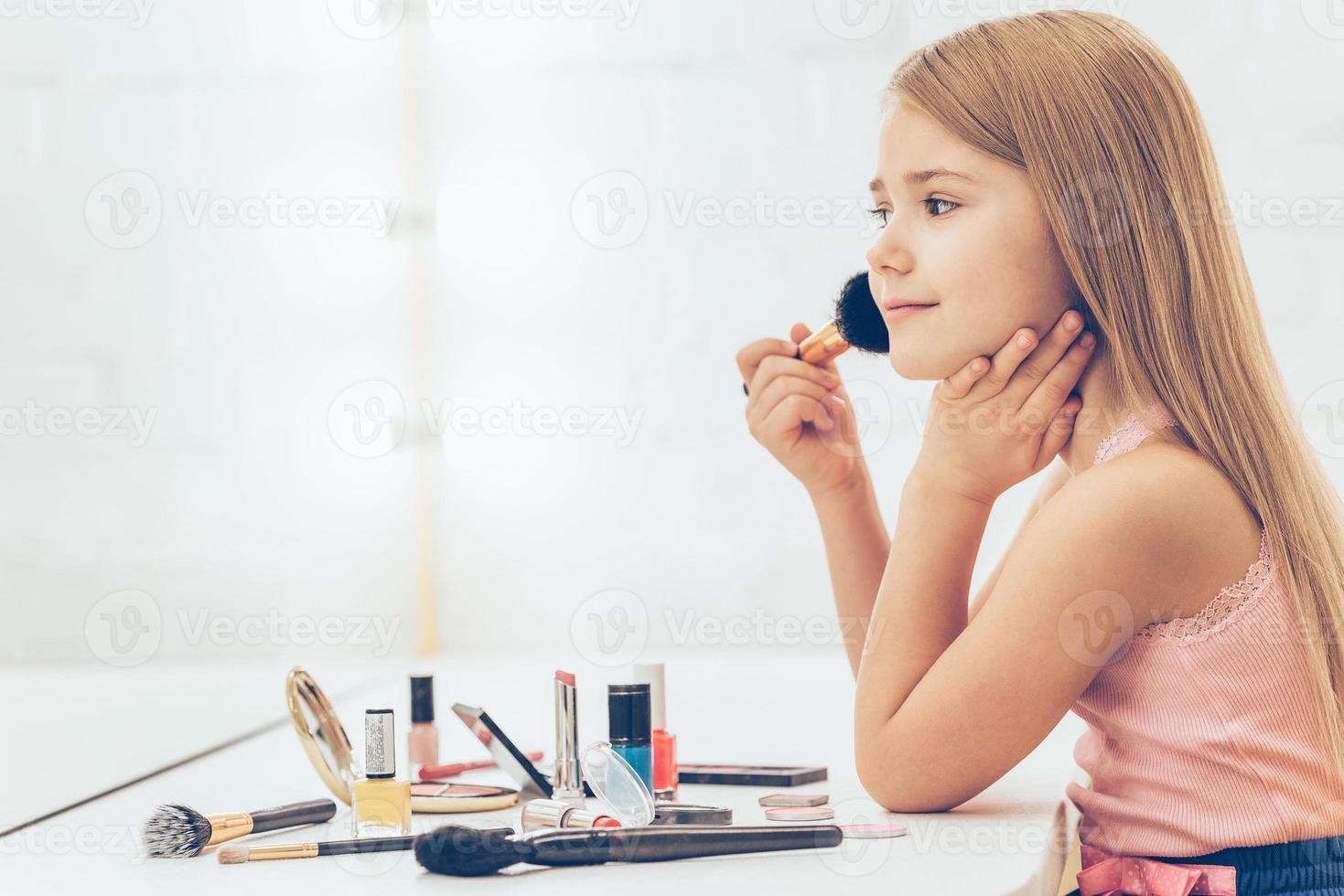 Adding some color to my cheeks. Side view of cheerful little girl applying make-up and looking at her reflection in mirror while sitting at the dressing table photo