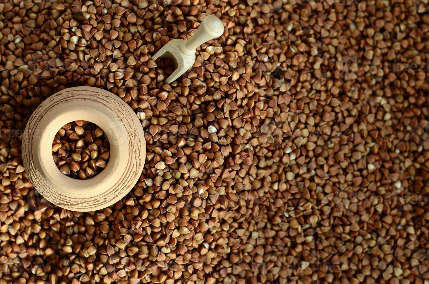 Background image of a large pile of buckwheat, in the middle of which lies a small jug and a wooden spatula for cereals photo