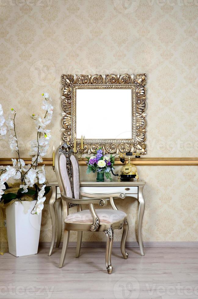 A fragment of a retro interior with a chair and table, on which is a telephone and a vase of flowers. Above the table hangs an empty picture frame for your photo
